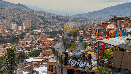 Large stone sculpture of a head overlooks a cityscape with hills, buildings, and a colorful sign reading "Medellin" in the foreground. People are gathered on a rooftop terrace.