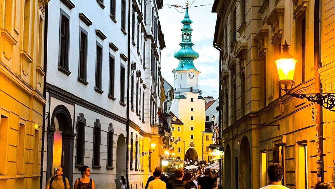 Narrow street in Bratislava lined with historical buildings, leading to a clock tower in the distance. People walk under warm streetlights at dusk.