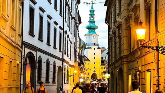 Narrow street in Bratislava lined with historical buildings, leading to a clock tower in the distance. People walk under warm streetlights at dusk.