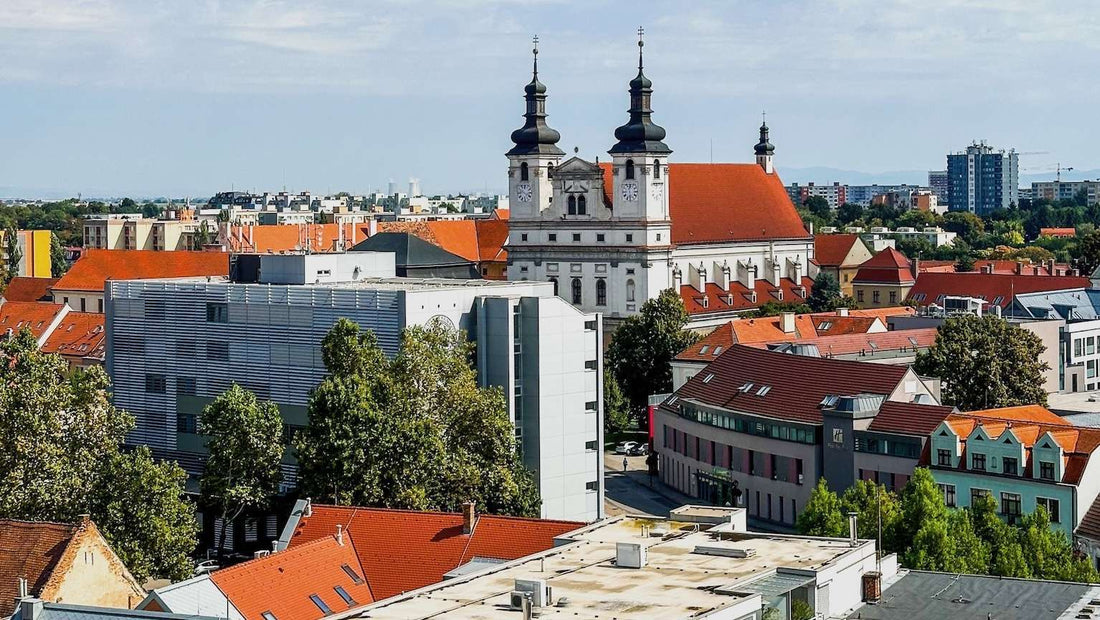 Aerial view of a Trnava, Slovakia cityscape featuring a baroque church with twin towers, surrounded by modern and traditional buildings with red-tiled roofs.
