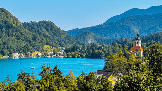 A scenic Lake Bled in Slovenia surrounded by forested hills, with small boats on the water and a church with a tall steeple on the right side, under a clear blue sky.
