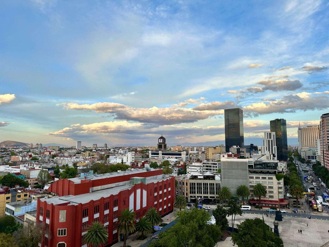 Cityscape view of Mexico City with modern skyscrapers and older red and white buildings, tree-lined streets, and a partly cloudy dusk sky—a vibrant backdrop to the city’s renowned food scene.