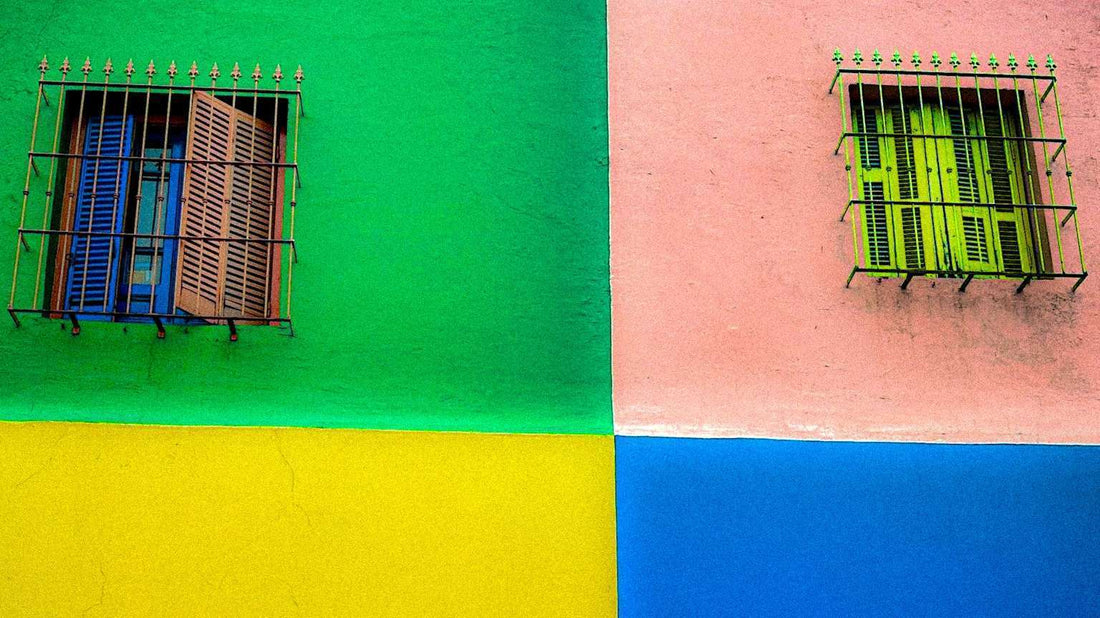 Two windows with metal bars against a vibrant, multicolored wall in Buenos Aires, Argentina divided into green, yellow, blue, and pink sections. The left window has wooden shutters; the right window has green shutters.
