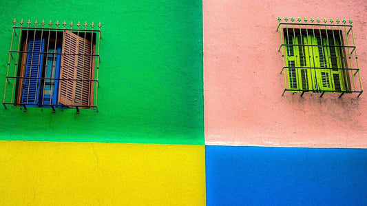 Two windows with metal bars against a vibrant, multicolored wall in Buenos Aires, Argentina divided into green, yellow, blue, and pink sections. The left window has wooden shutters; the right window has green shutters.
