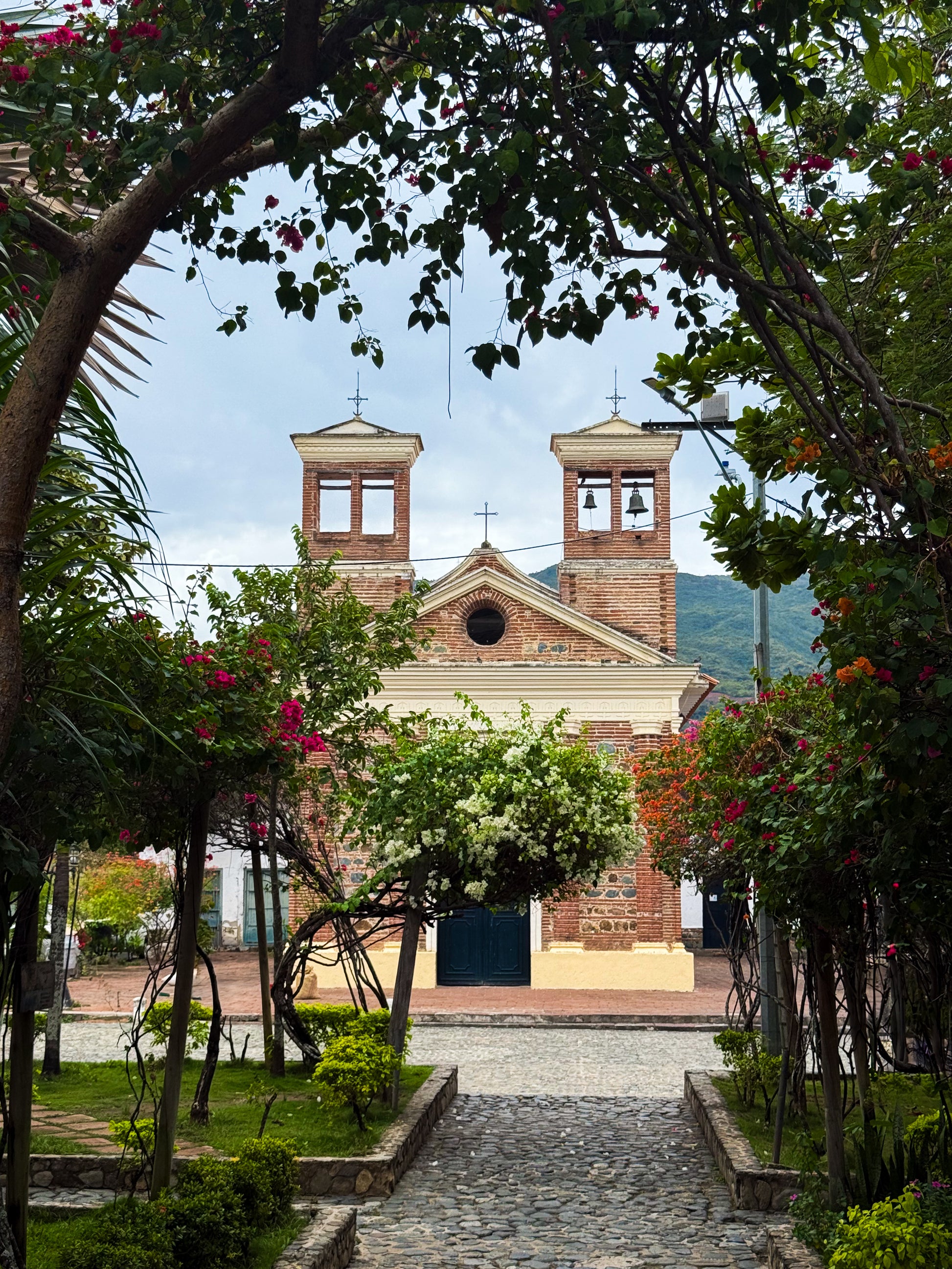 Charming Church - Fine Art Print by Leslie Brashear Photography features a brick church with twin bell towers framed by flowering plants and greenery along a cobblestone path, capturing classic Colombian architecture.