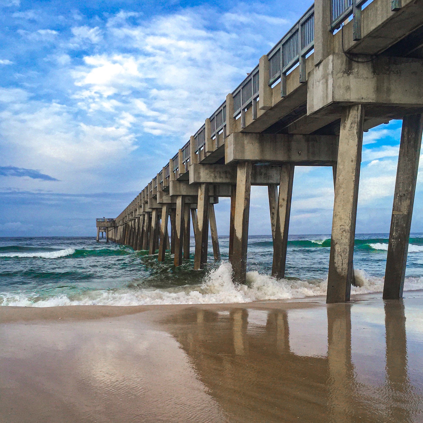 Pier Reflection - Fine Art Print captures a concrete pier stretching over ocean waves, with wet sand reflecting the scene below a partly cloudy sky, showcasing the tranquility and artistry of coastal photography.