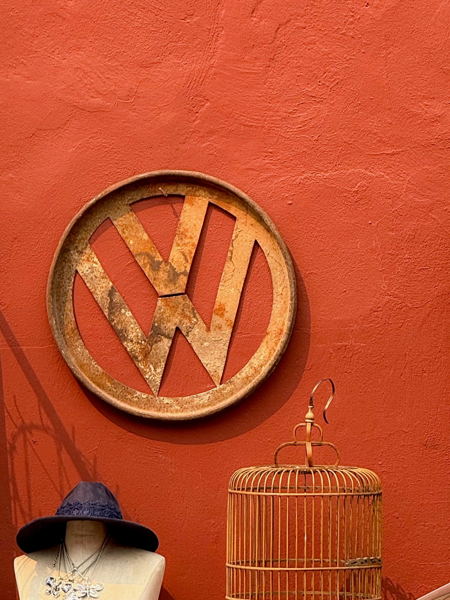 The VW Sign for Sale - Fine Art Print by Leslie Brashear Photography features a vintage Volkswagen logo displayed on a textured red wall above a mannequin with a hat and a decorative birdcage.