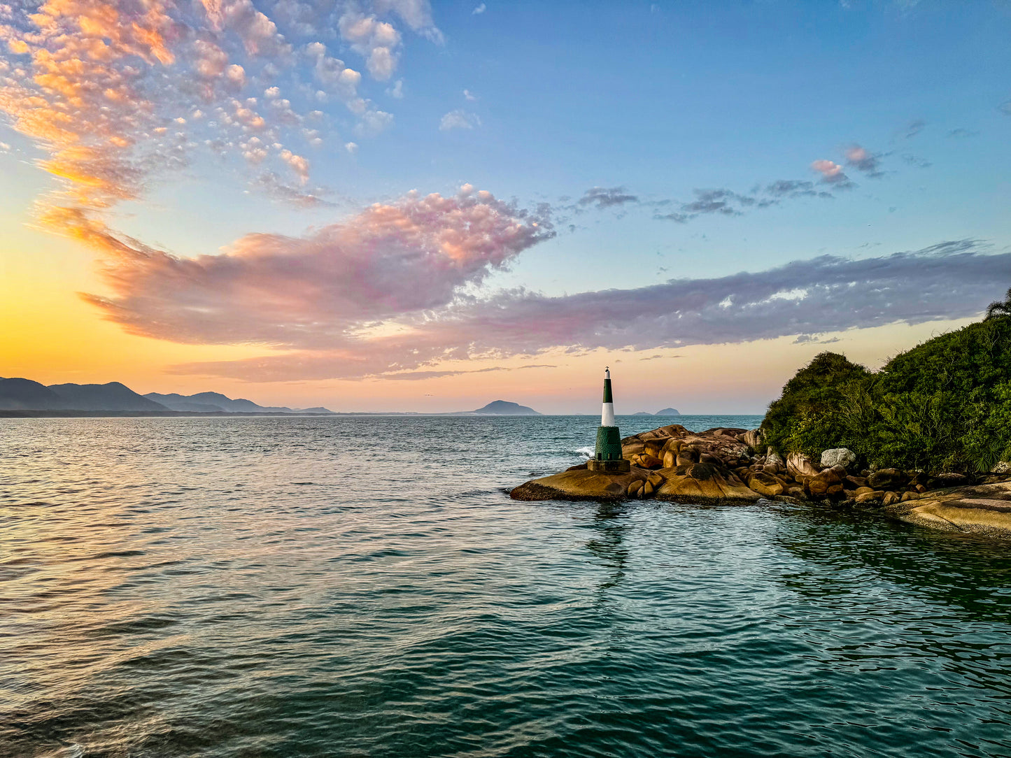 Coastal Sunset - Fine Art Print depicts a small lighthouse on a rocky shore at sunset, evoking the beauty of a Brazilian seascape with distant mountains and dramatic clouds.