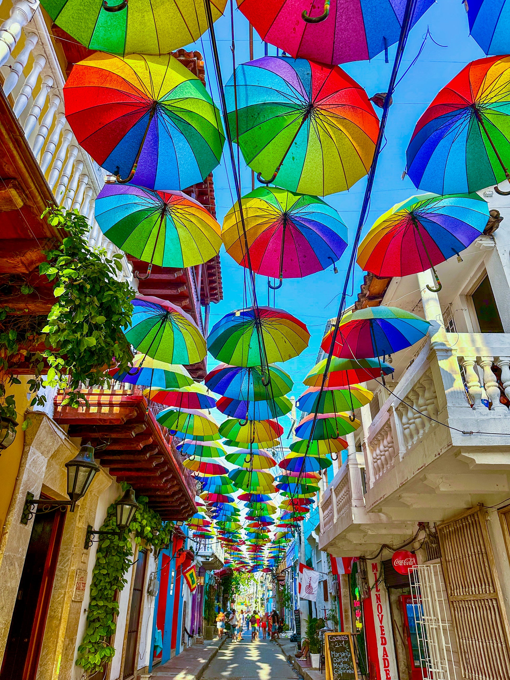 In this Leslie Brashear Photography print, “Umbrellas - Fine Art Print,” colonial architecture lines a narrow Cartagena street as colorful umbrellas hang overhead, forming a lively canopy beneath the clear blue sky.