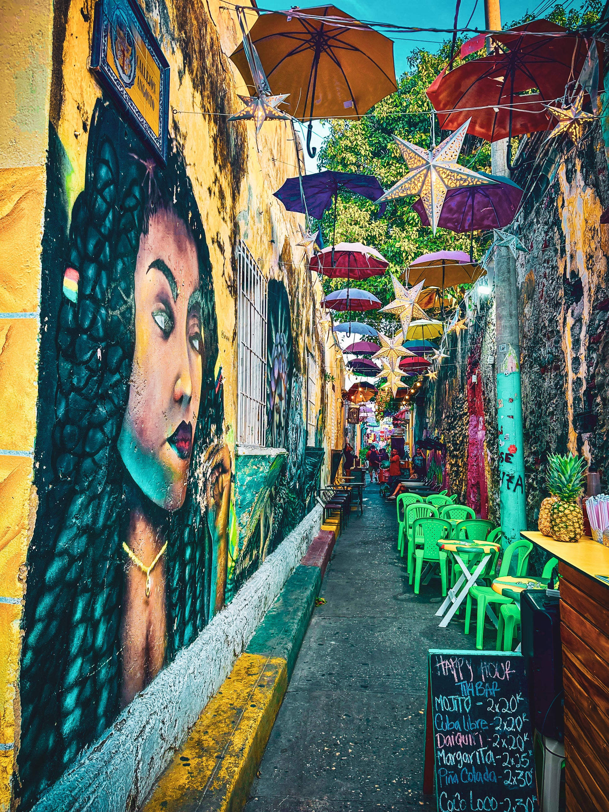 Getsemani Alley - Fine Art Print by Leslie Brashear Photography features a vibrant Cartagena mural, colorful umbrella street art, green chairs, tables, and a chalkboard menu with drink prices.