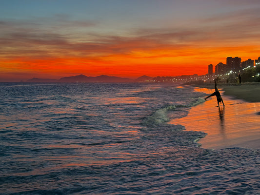 Sunset Cartwheels at Ipanema Beach - Fine Art Print by Leslie Brashear Photography captures a person cartwheeling at sunset on Ipanema Beach, with city lights, ocean waves, and Rio's mountains in the background.